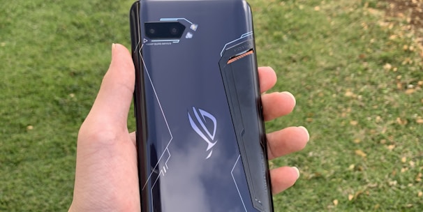 A hand holding a sleek, dark-colored smartphone with geometric patterns and the text 'Tencent Games' on the back cover. The background features a grassy area with some trees or bushes on the side.